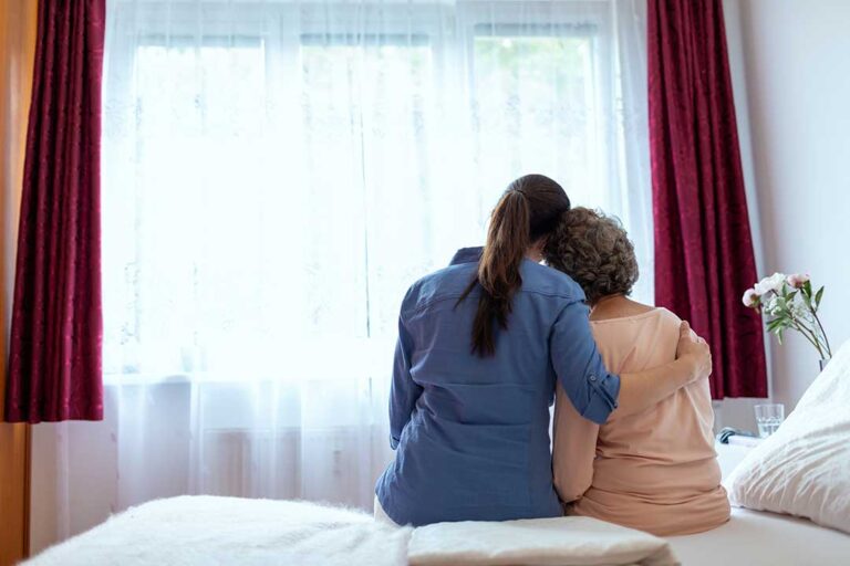 An elderly woman and a younger visitor holding hands and talking in a bright common room with personal items visible nearby.
